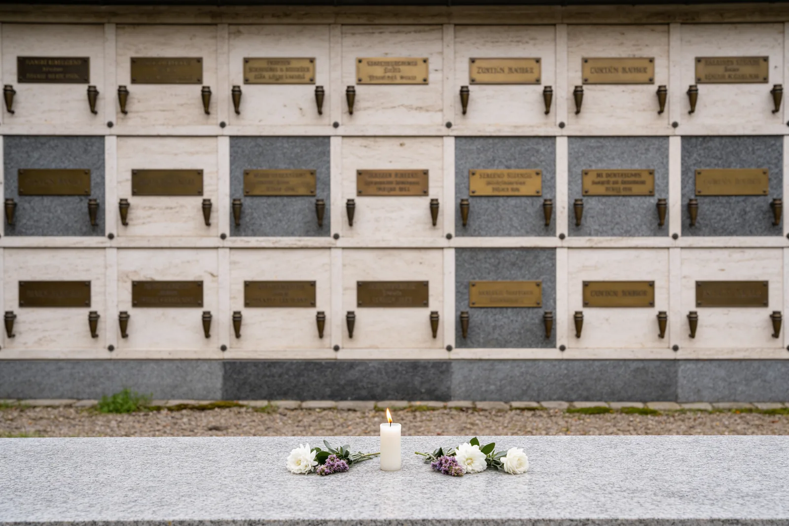 Gepflegte Urnenwand (Kolumbarium) auf einem Friedhof mit reihenweise angeordneten Nischen und beschrifteten Verschlussplatten, davor eine Kerze und Blumen – Sinnbild für eine Kolumbarium-Bestattung